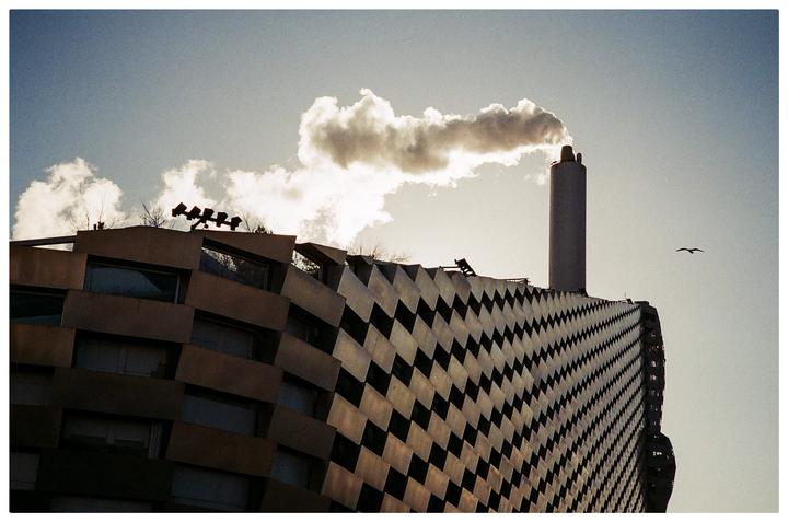 A tall smokestack emits a large cloud of smoke next to a building with a patterned facade.
