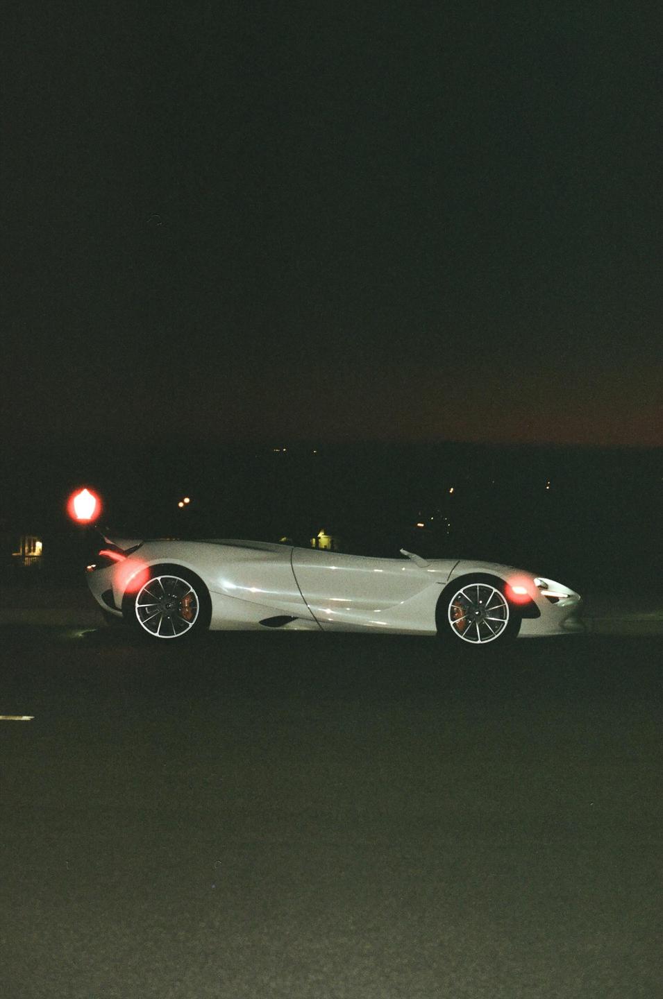 A white sports car with its lights on is parked on a dark road at night.