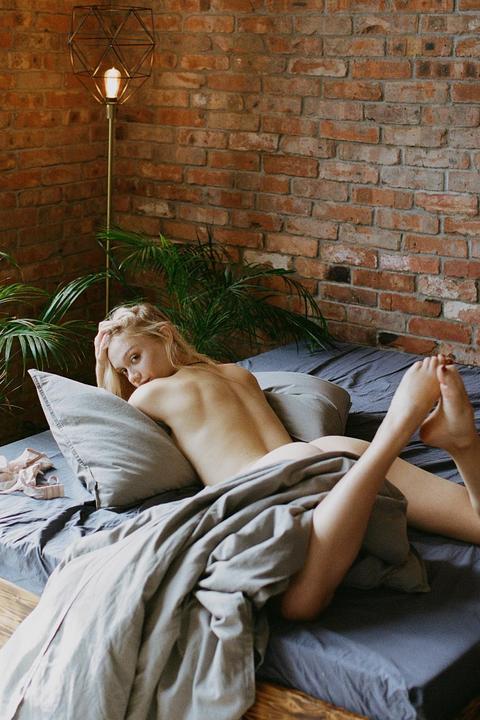 A woman lies on a bed with a sheet covering her lower body, looking over her shoulder at the viewer.