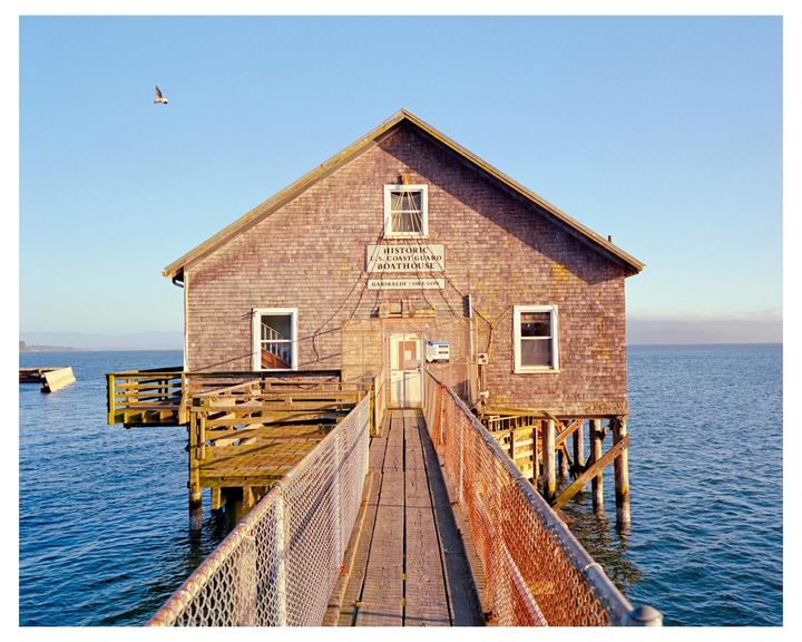 A wooden boathouse stands over blue water with a bird flying in the clear sky.