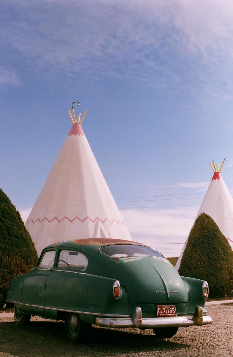 A green vintage car is parked in front of two white teepees with pink and red trim under a blue sky.