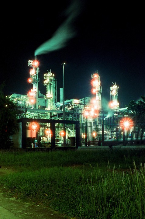 A large industrial facility with smoke rising from a smokestack is brightly lit at night, with tall grass in the foreground.