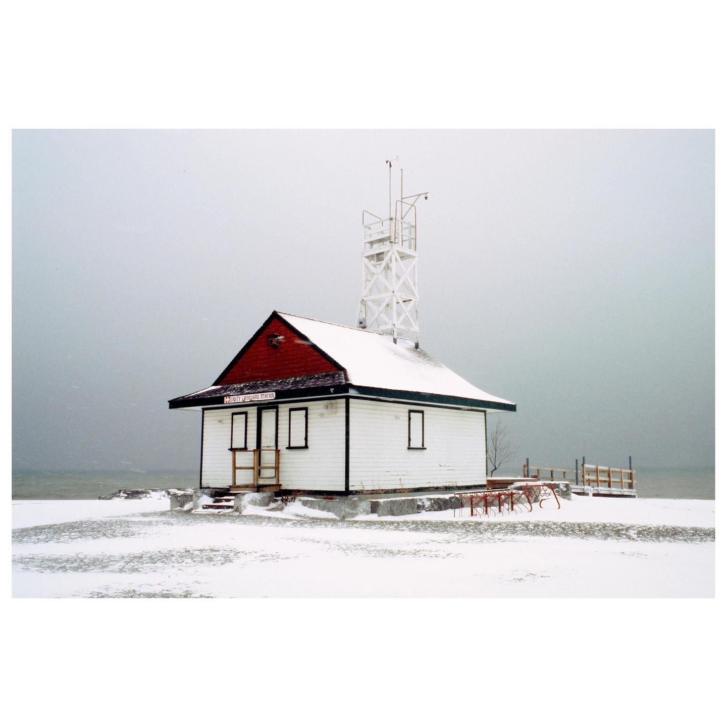 A small white building with a red roof and a tall white tower stands in a snow-covered landscape next to a body of water.