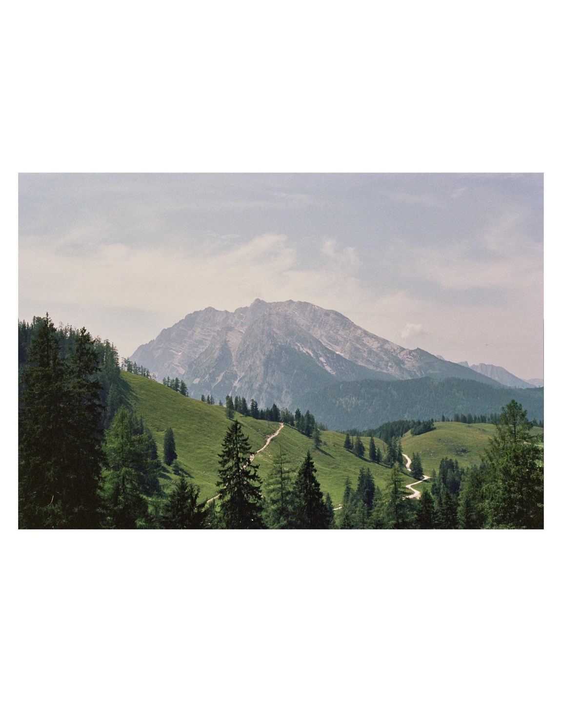 A winding path leads through a green, forested valley towards a large mountain under a cloudy sky.