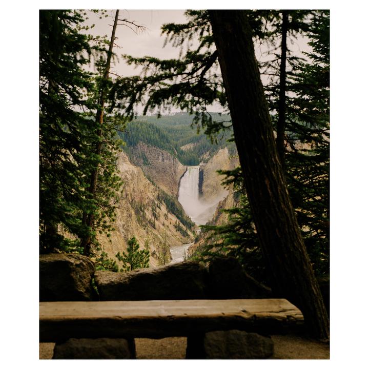 A waterfall cascades into a canyon, framed by pine trees and a stone wall.