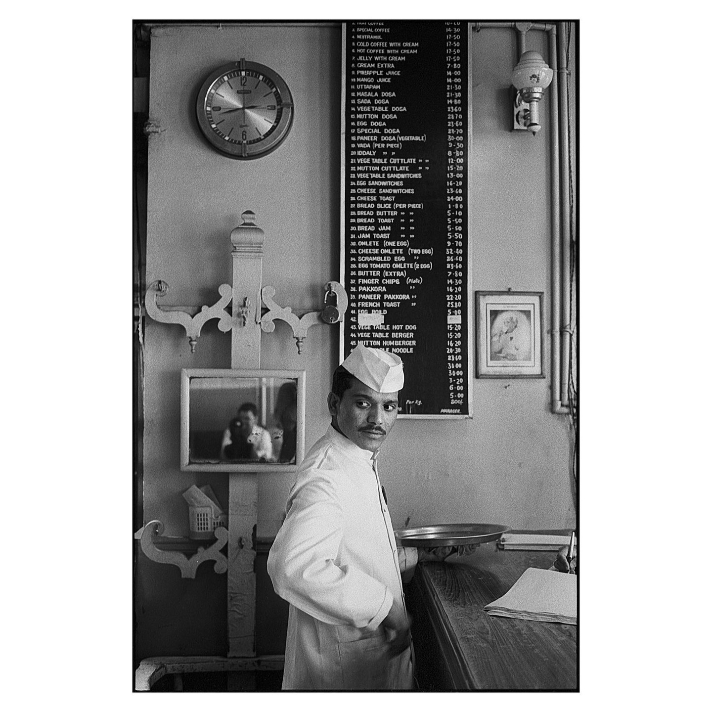 A man in a uniform stands behind a counter in a coffee house with a menu board and a clock on the wall.