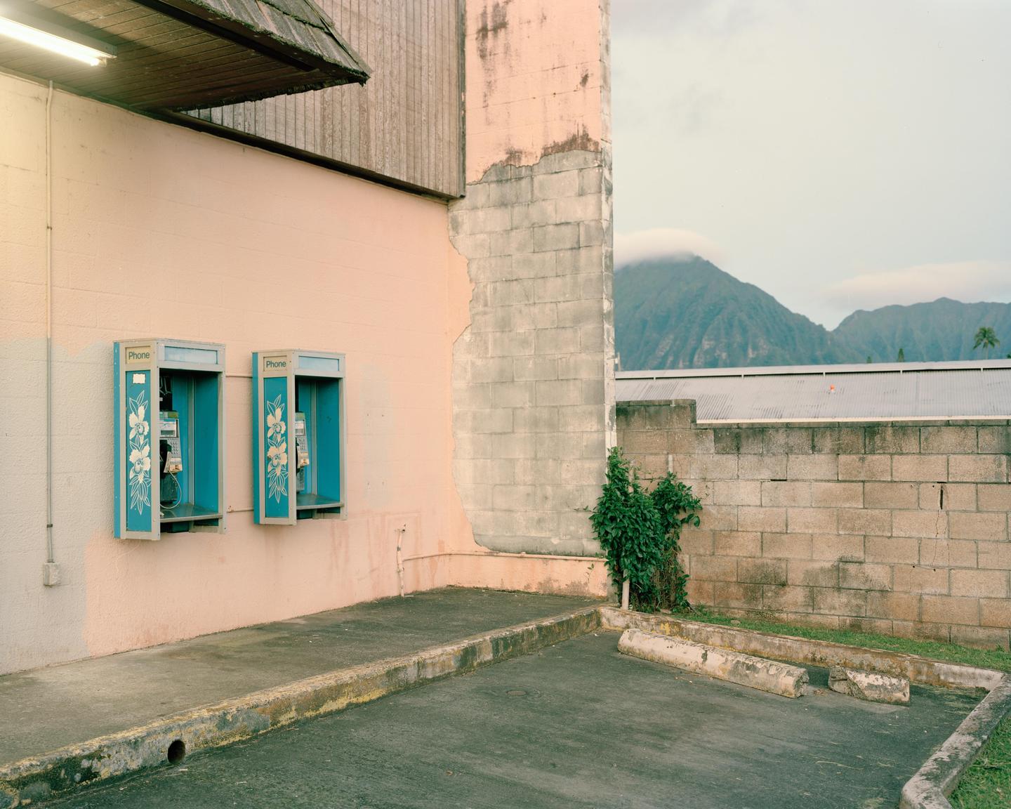 Two payphones are mounted on a pink wall next to a parking lot with a mountain in the background.