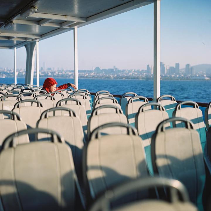 A person in a red jacket sits on a ferry looking out at a city skyline across the water.