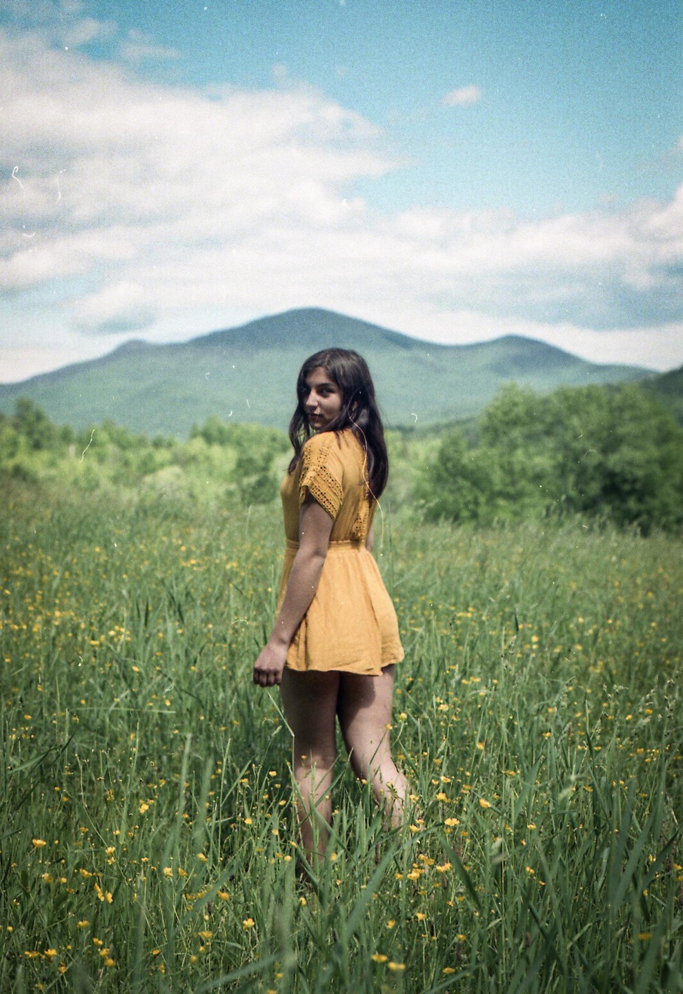 A woman in a yellow dress stands in a field of tall grass and yellow flowers with mountains in the background.