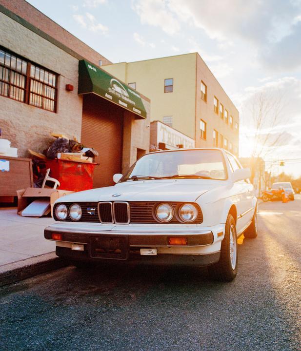 A white car is parked on a street with buildings and a dumpster in the background, illuminated by the setting sun.