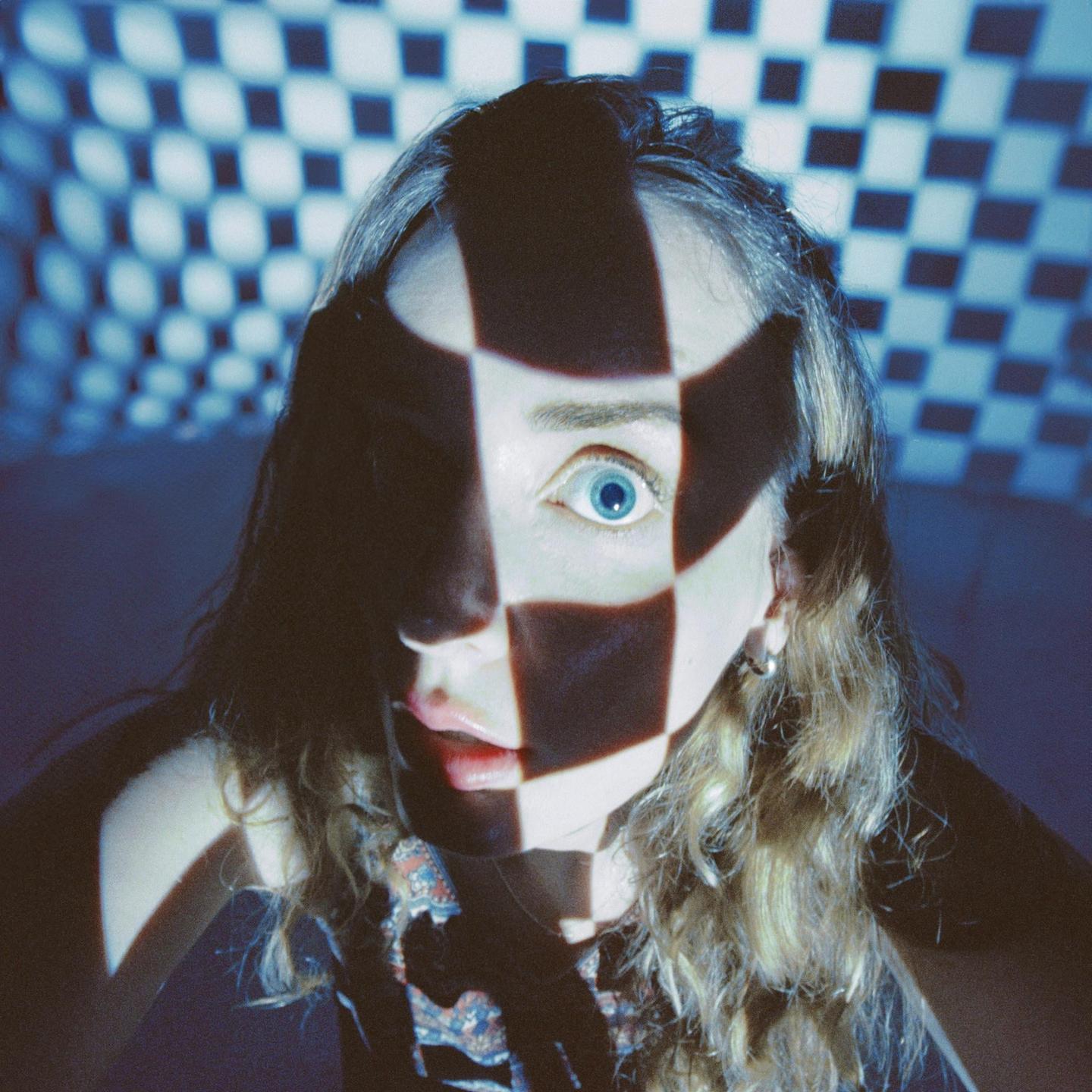 A woman with blue eyes looks forward, with a checkerboard pattern projected onto her face and the wall behind her.