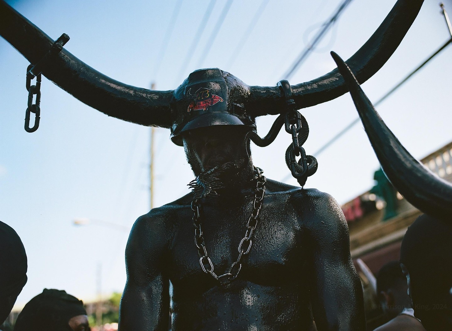 A man covered in black paint and wearing a horned helmet and chains stands under a bright sky.
