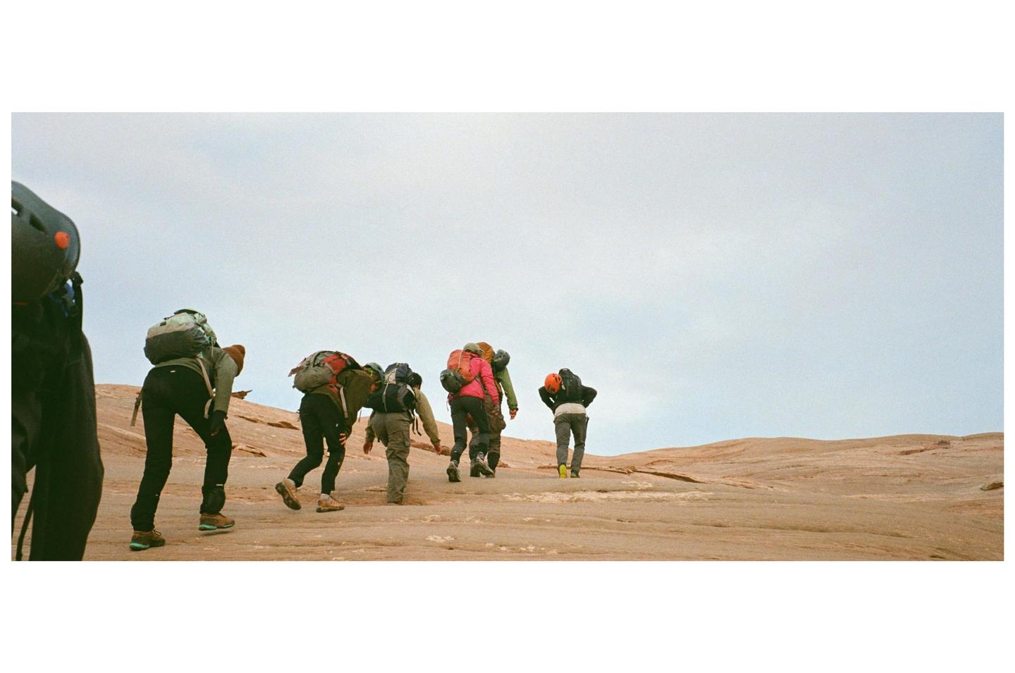 A group of people with backpacks hike up a rocky, barren hill under a light sky.
