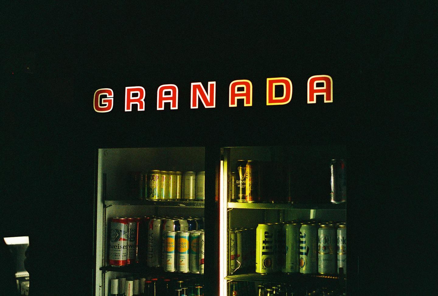 A red neon sign spelling "GRANADA" is visible above a refrigerated display case filled with beverage cans.