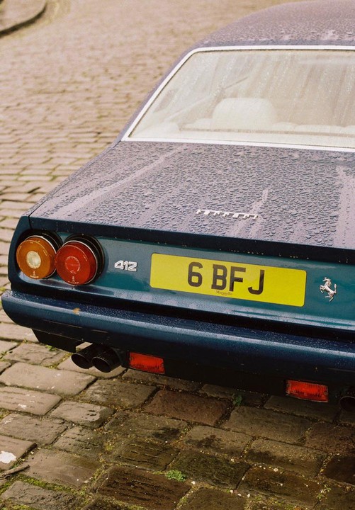 The rear of a dark blue Ferrari 412 is covered in raindrops and parked on a cobblestone street.