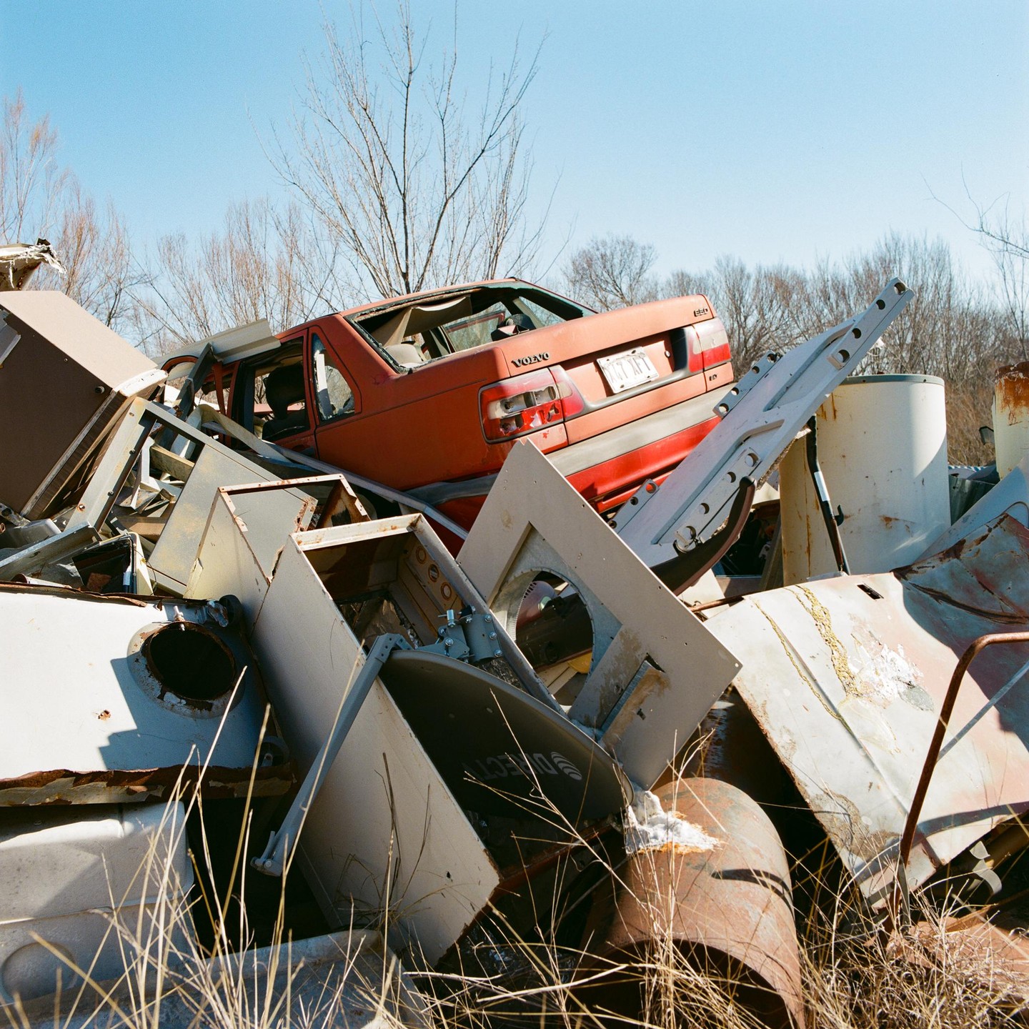 A red Volvo is visible in a junkyard, surrounded by various metal scraps and dry grass under a clear sky.