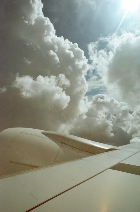 An airplane wing and engine are visible against a sky filled with large, fluffy clouds and a bright sun.