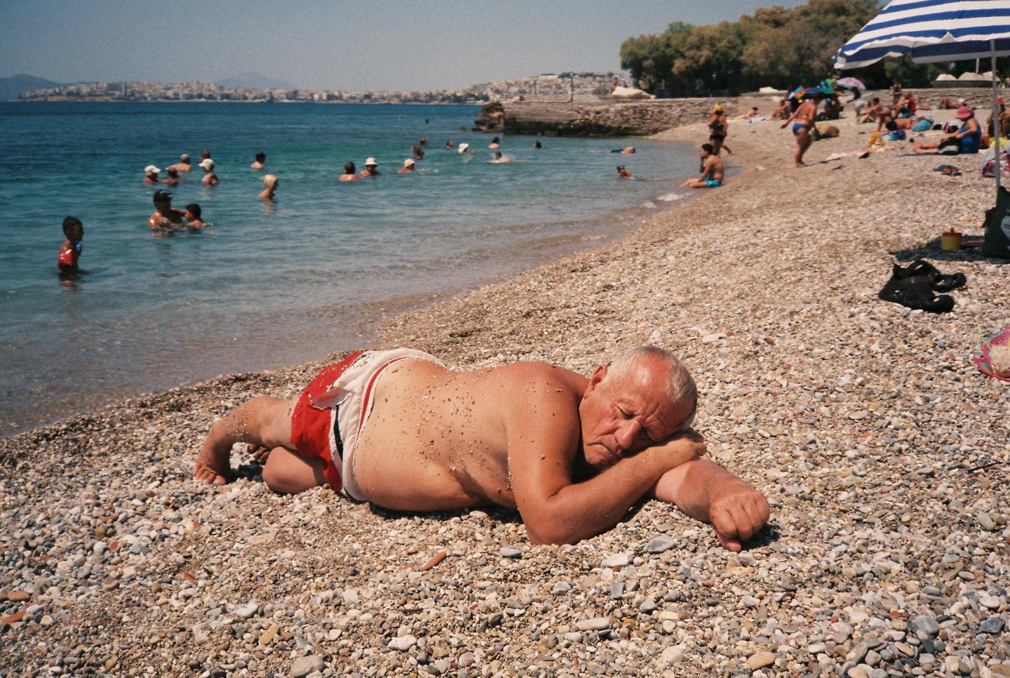 A man in red swim trunks sleeps on a pebble beach with people swimming in the sea and a city in the background.