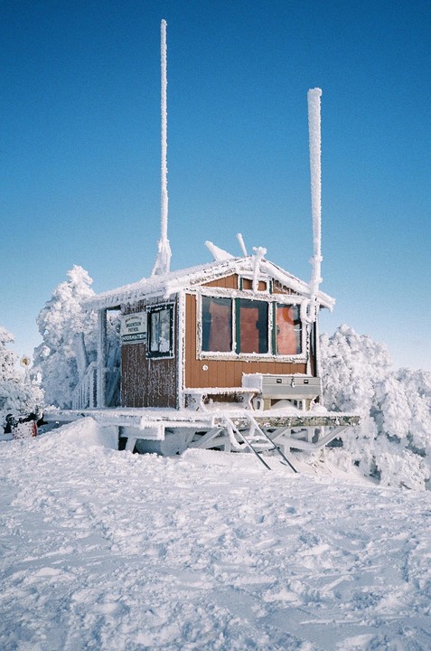 A small wooden cabin covered in snow and ice stands on a snowy mountain under a clear blue sky.