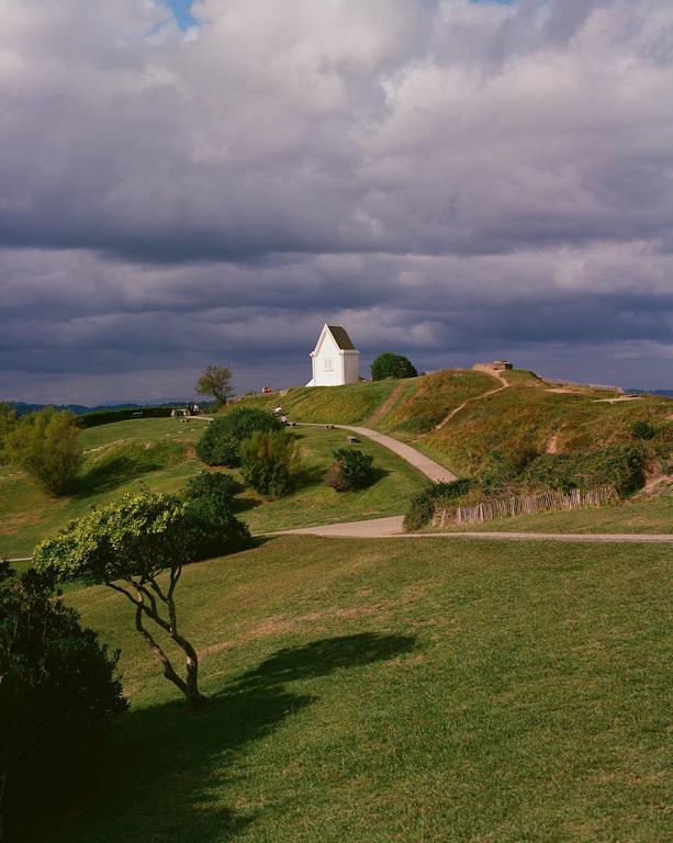 A small white building sits atop a grassy hill under a cloudy sky, with winding paths and trees in the foreground.