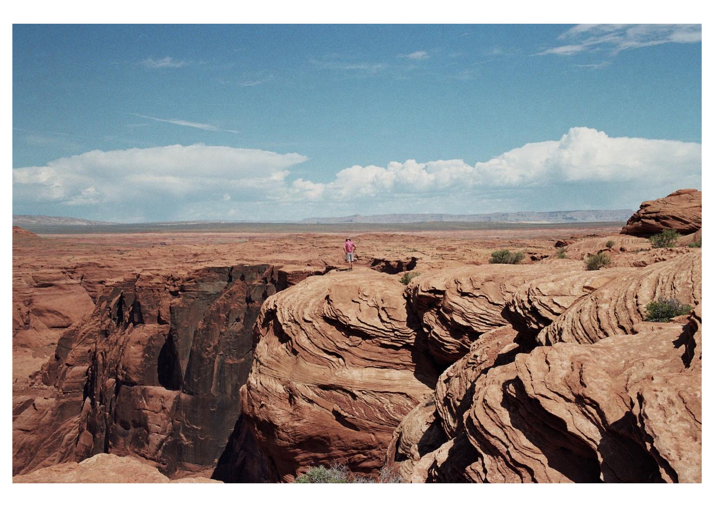 A man stands on a cliff overlooking a vast desert landscape under a blue sky with clouds.