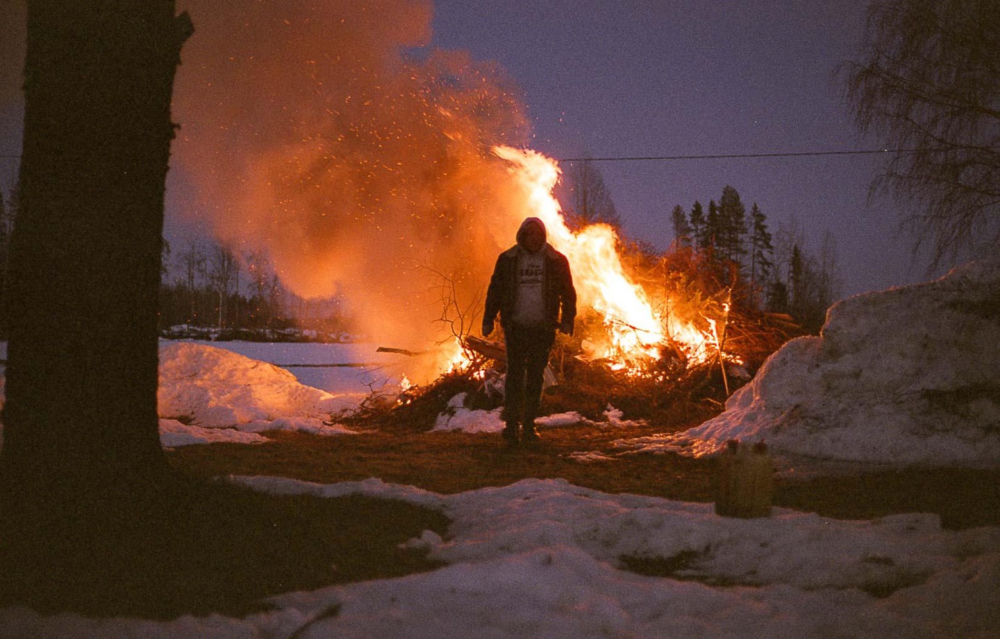 A person stands in front of a large bonfire with snow on the ground and trees in the background at dusk.