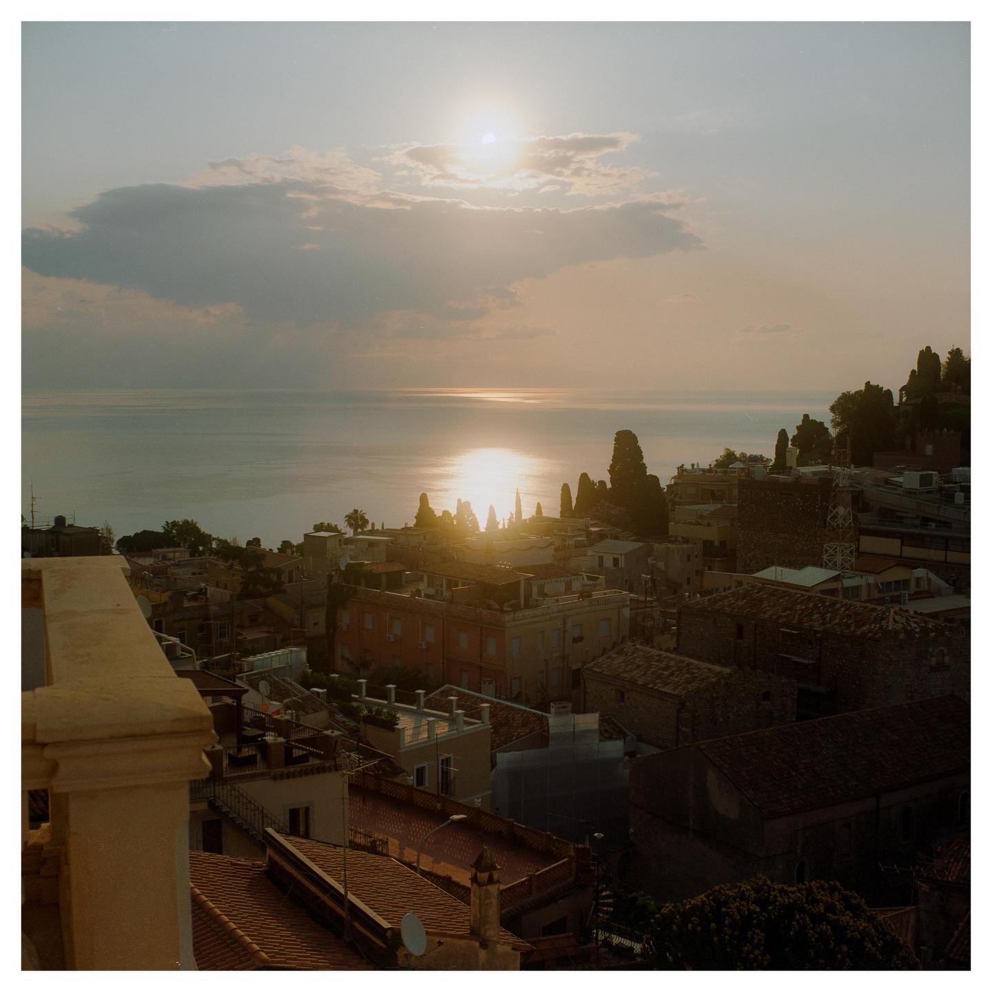 The sun sets over a town on a hill overlooking the sea, with a bright reflection on the water.