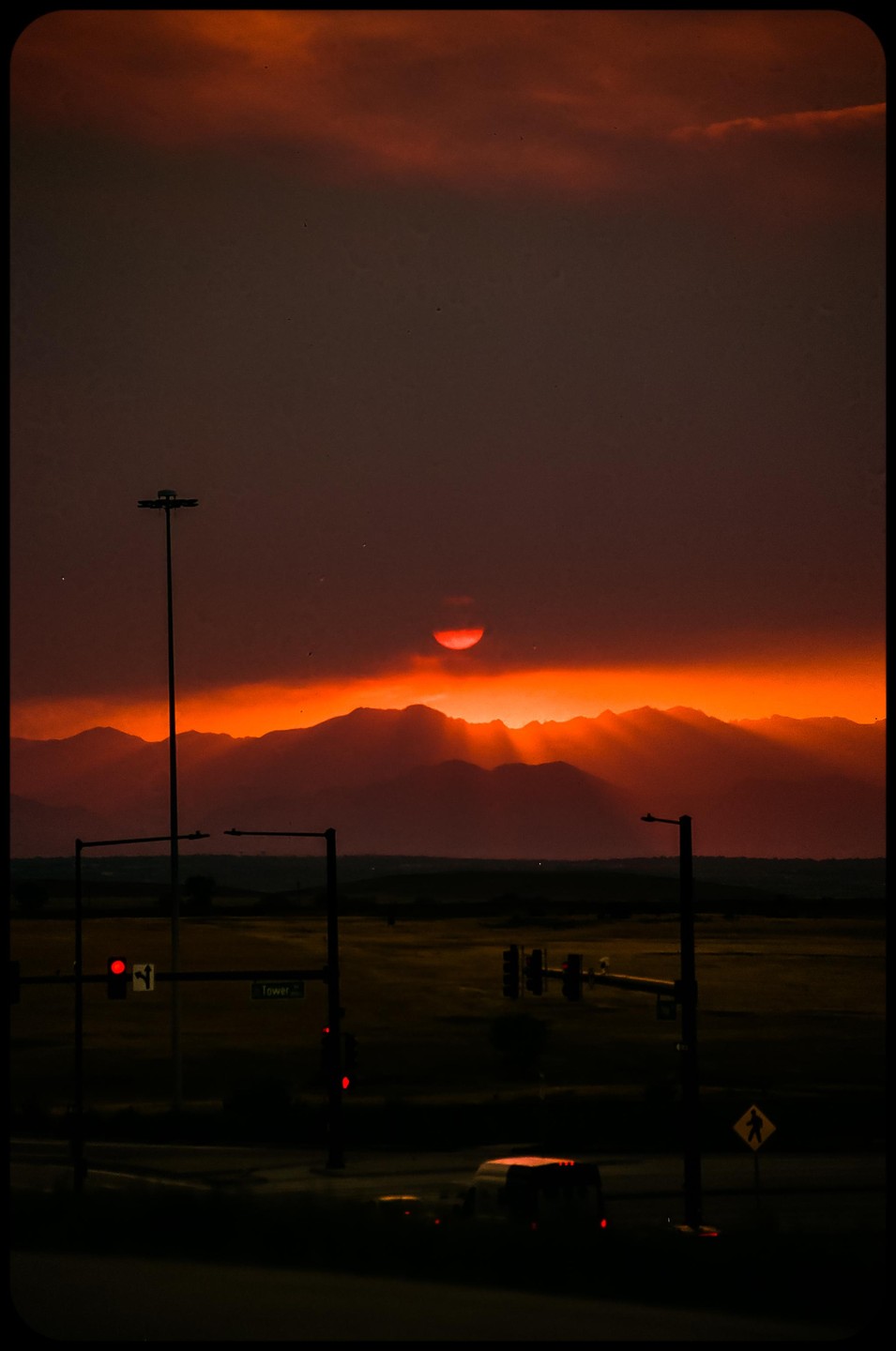 The sun sets behind mountains, casting an orange glow over a dark landscape with streetlights and traffic lights.