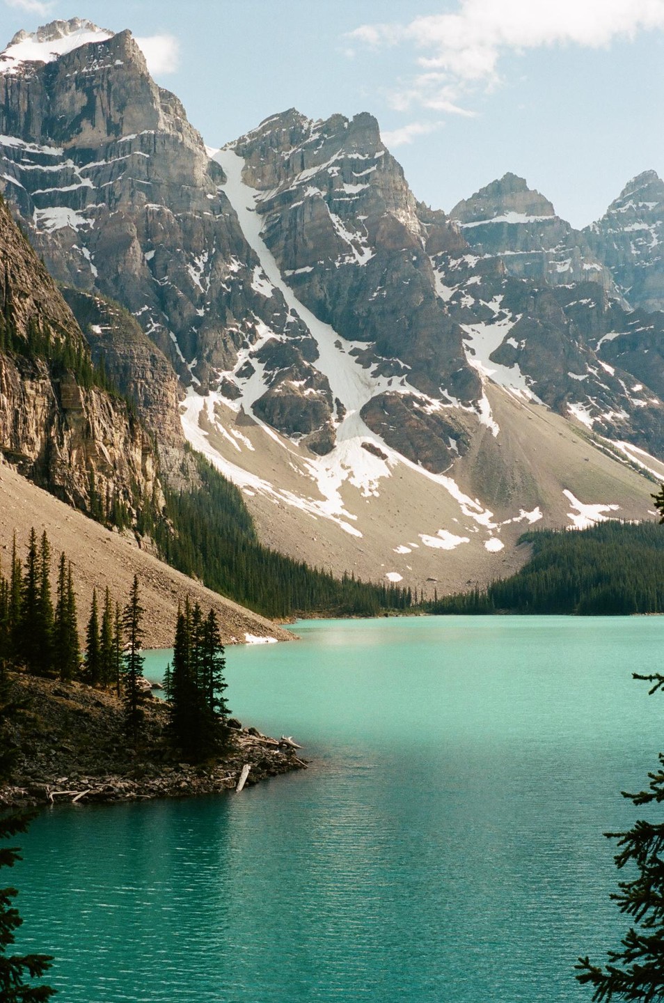 A turquoise lake is surrounded by snow-capped mountains and a pine forest.
