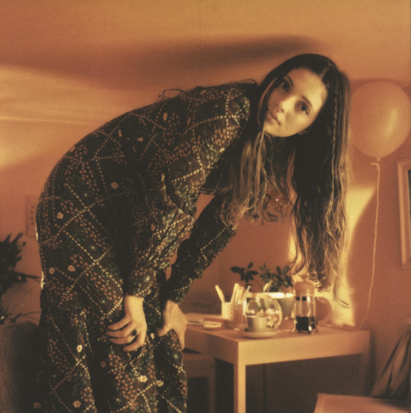 A woman in a patterned dress bends over a table with a coffee pot and a balloon in the background.