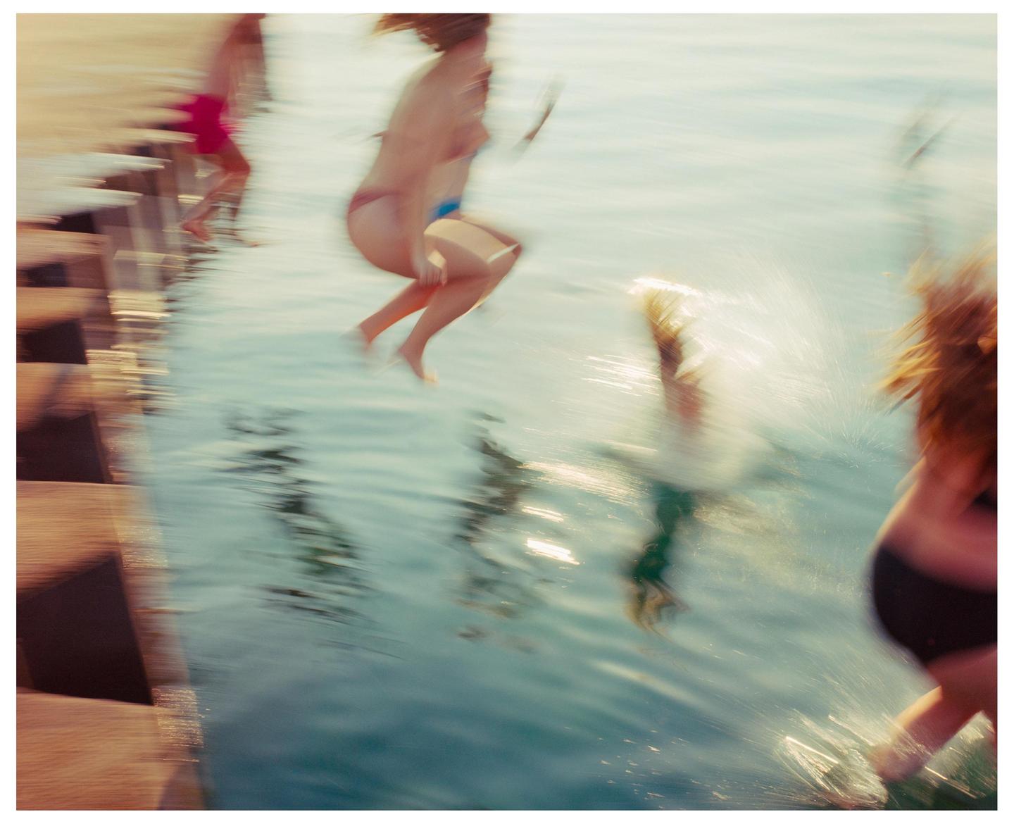 Several people jump into the water from a wooden dock, creating splashes.