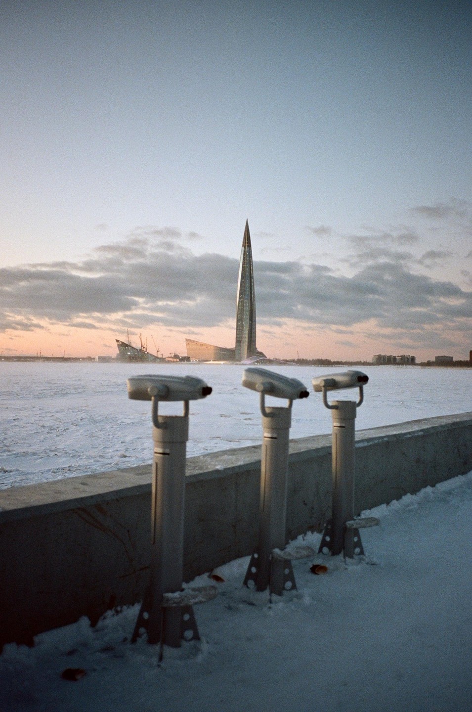 Three coin-operated binoculars overlook a frozen body of water with a modern skyscraper and other buildings in the distance under a cloudy sky.