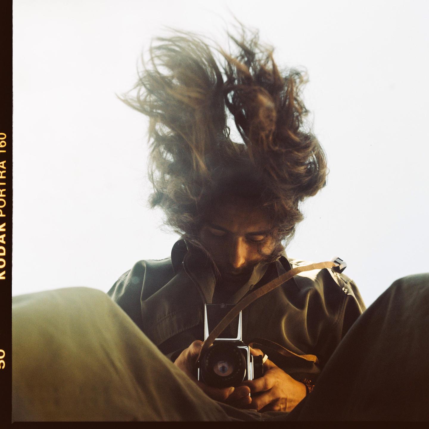 A man with long hair flying upwards holds a camera and looks down at it.