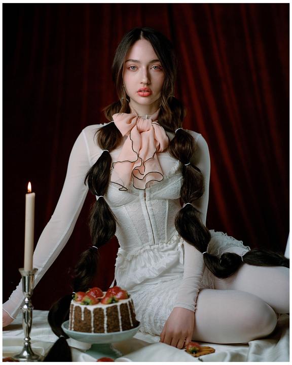 A woman with long braided hair and a large pink bow sits next to a chocolate cake with strawberries and a lit candle.