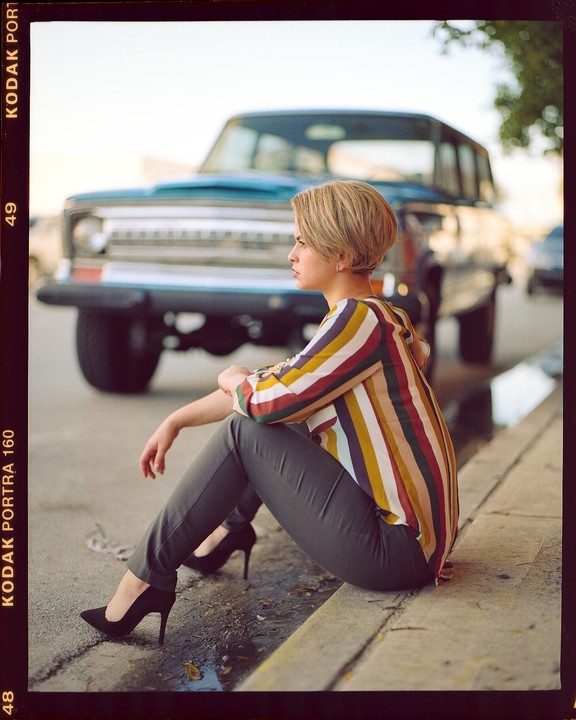 A woman with short hair and a striped shirt sits on a curb next to a jeep.