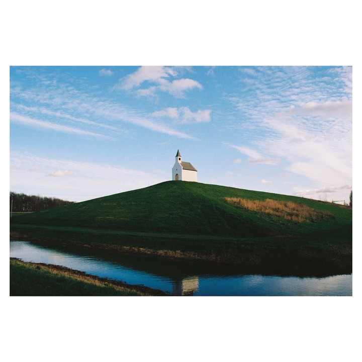 A small white chapel with a dark steeple sits atop a grassy hill next to a narrow body of water under a blue sky with wispy clouds.