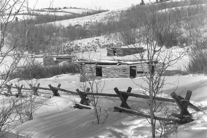 Three wooden cabins and a split-rail fence are visible in a snowy landscape with hills and trees in the background.