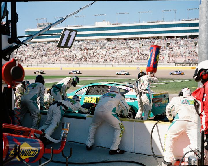 A pit crew services a race car on a track with a grandstand full of spectators in the background.