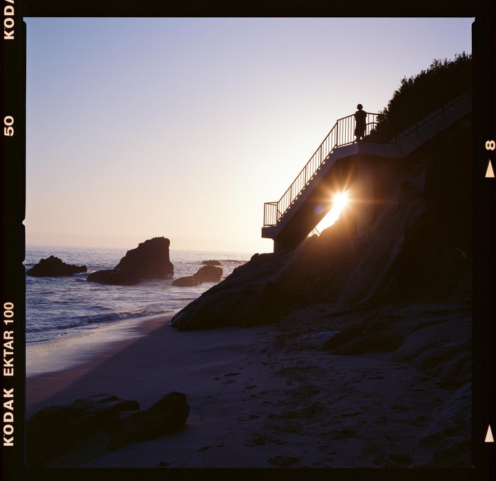 A person stands on a staircase overlooking a beach with rocks and ocean waves at sunset.