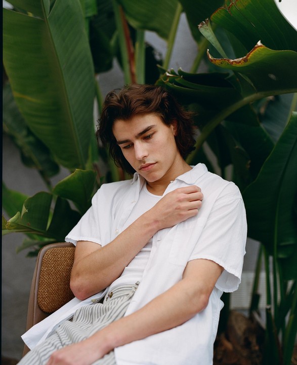 A young man with long hair sits in a chair in front of large green leaves.