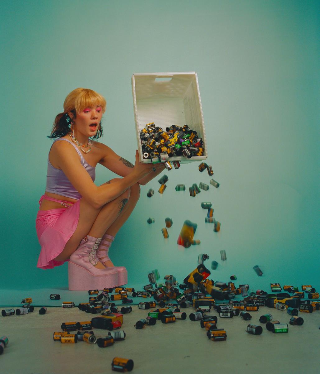 A woman in a pink skirt and platform shoes crouches while pouring film canisters from a white basket onto the floor.