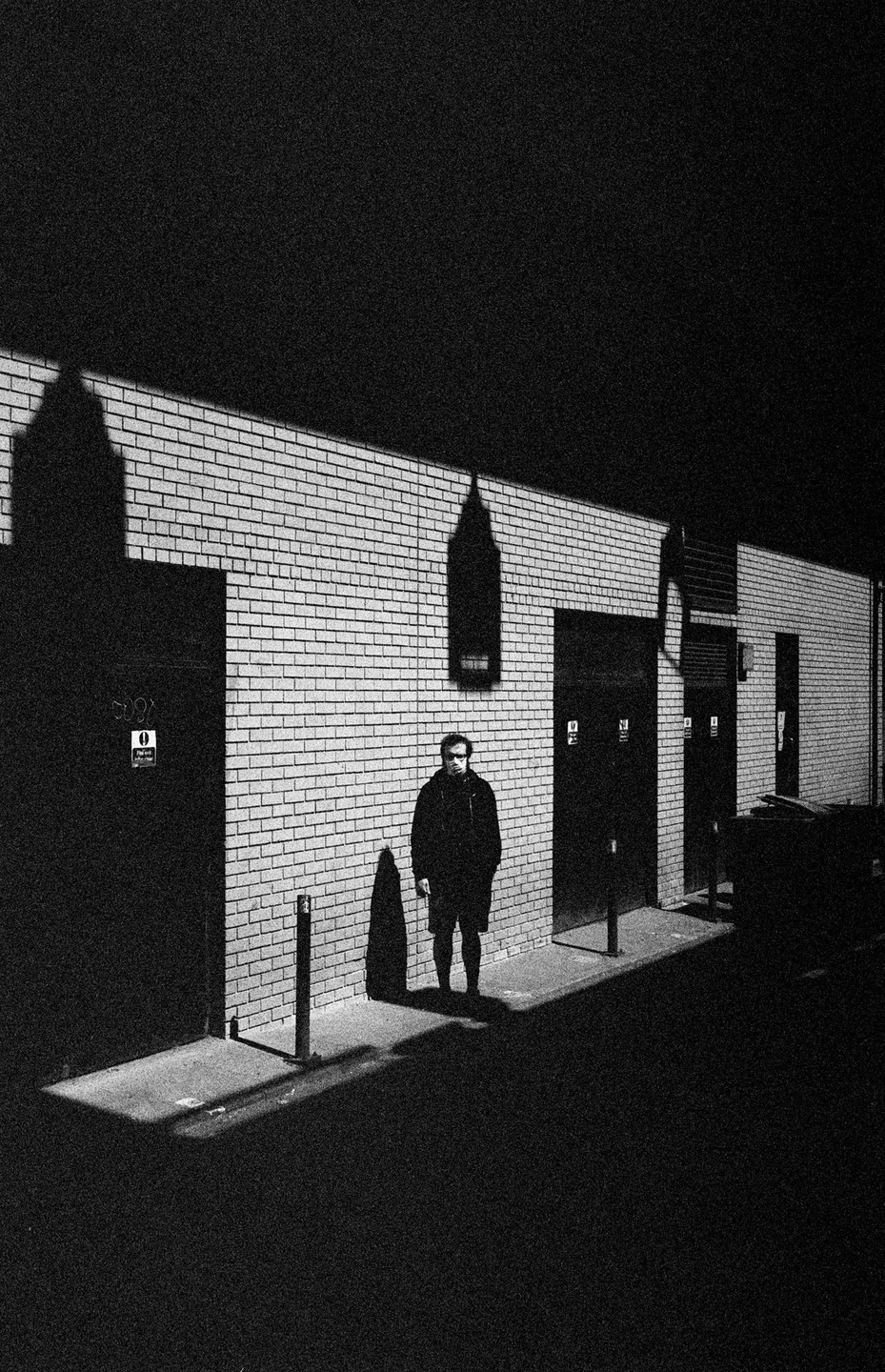 A man stands in an alley next to a brick wall with shadows cast upon it at night.