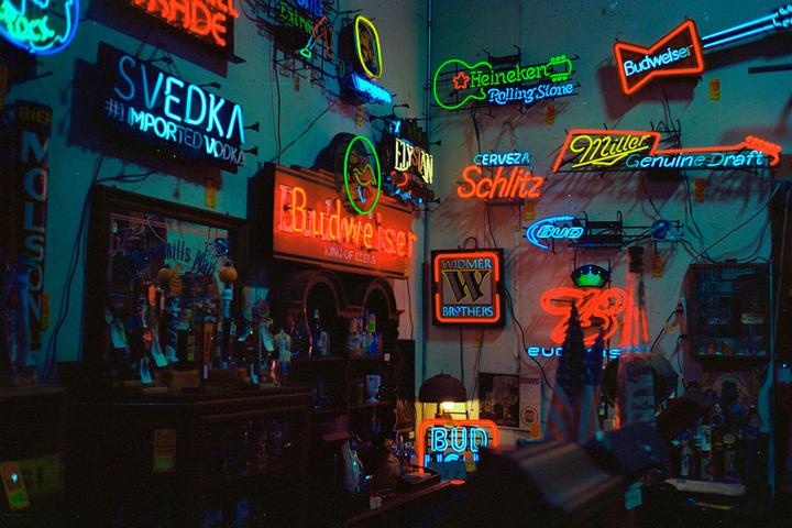 A bar interior is illuminated by numerous neon signs for various alcohol brands.