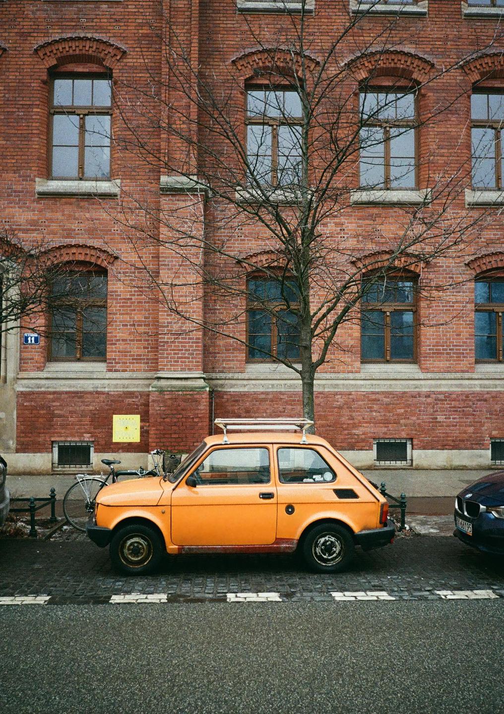 An orange car is parked on a street next to a brick building with a bare tree in front of it.