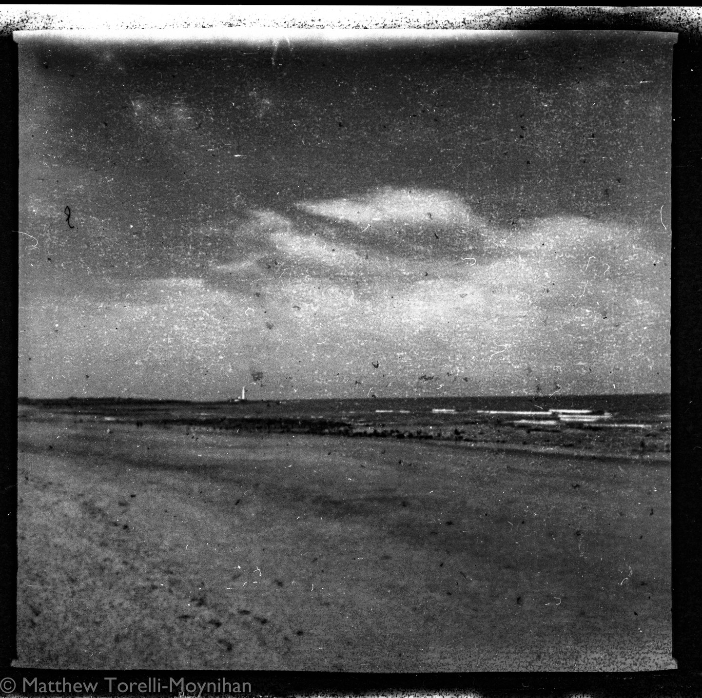 A black and white landscape shows a beach with a lighthouse in the distance under a cloudy sky.