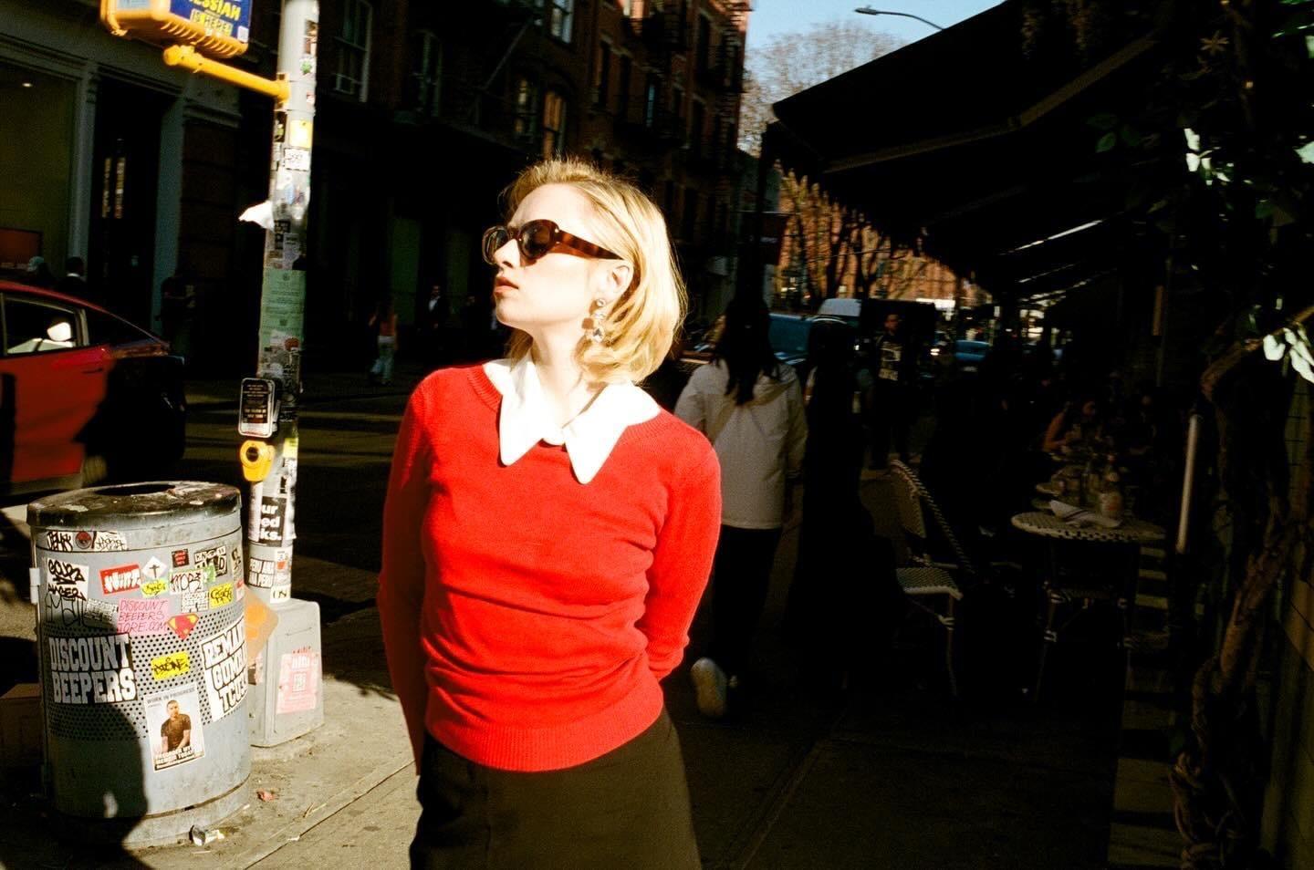 A woman in sunglasses and a red sweater stands on a sunlit street next to a trash can covered in stickers.