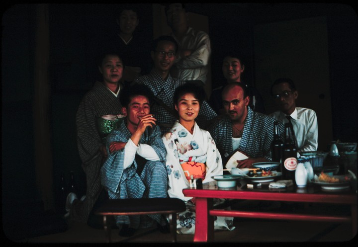 A group of people, some wearing kimonos, are gathered around a low table with food and drinks.