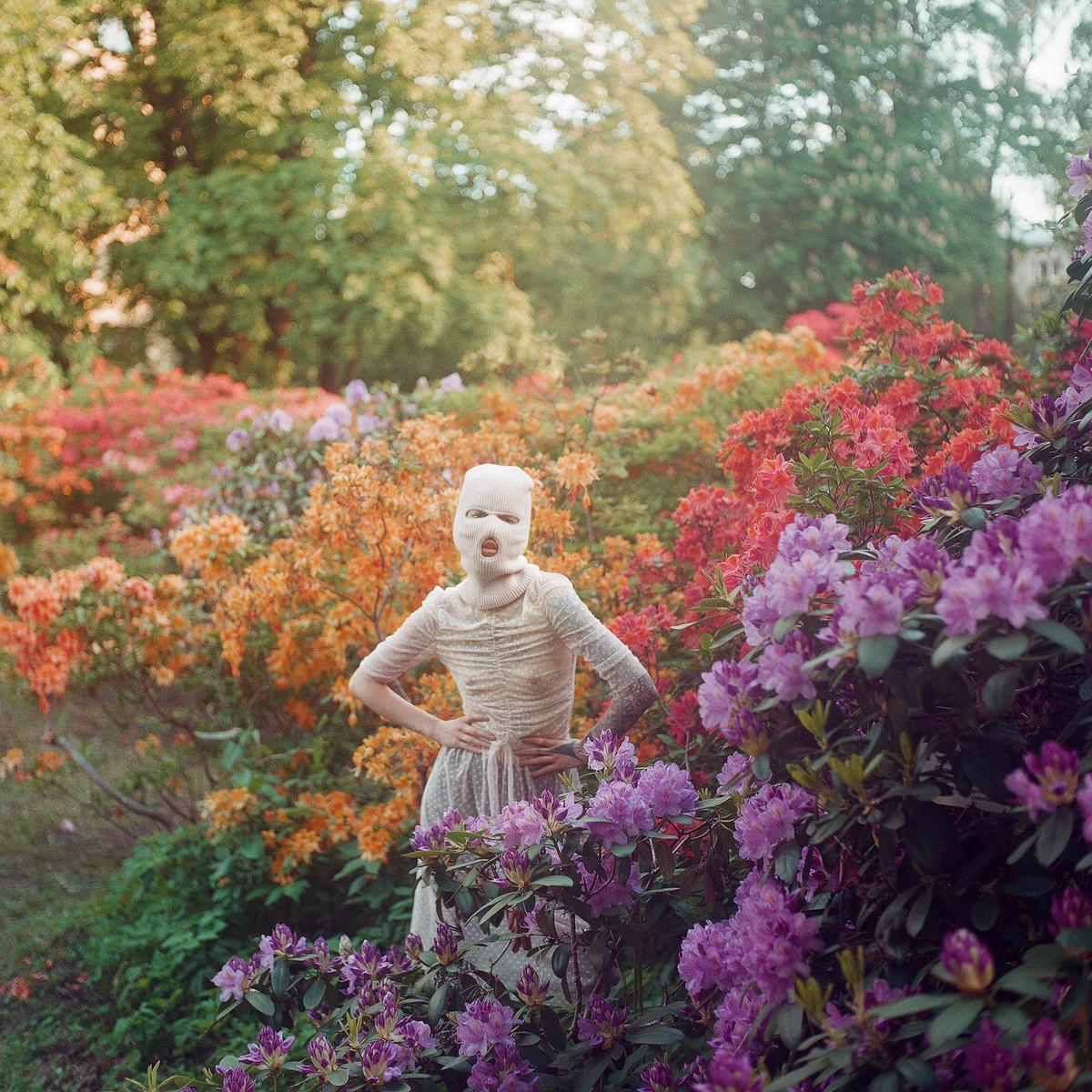 A person wearing a balaclava and a dress stands in a garden filled with colorful flowering bushes.