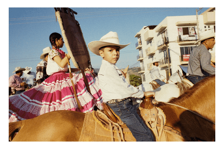 A boy in a cowboy hat sits on a horse next to a woman in a pink dress, with other people on horseback and buildings in the background.