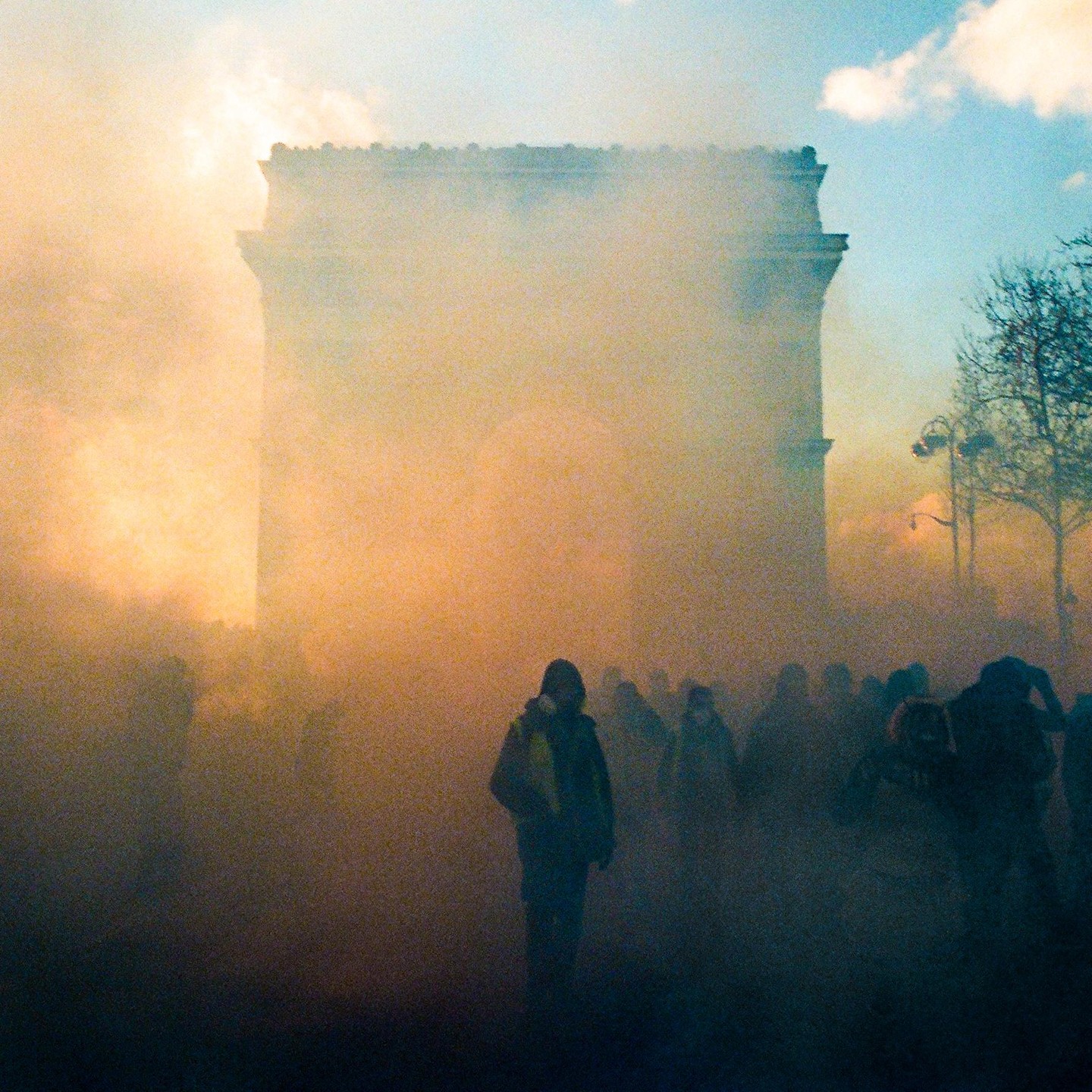 A crowd of people is shrouded in yellow smoke in front of the Arc de Triomphe.
